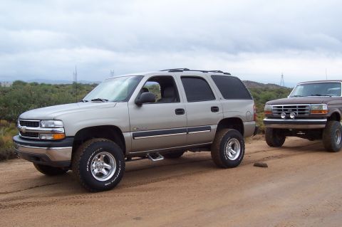 1992 Ford Bronco - Two SUVs are parked on a dirt road with scrub brush and hills in the background under a cloudy sky.