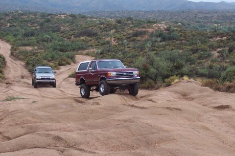1992 Ford Bronco - Two SUVs, a Ford Bronco and a Chevrolet Trailblazer, navigate a sandy off-road path with hills and sparse vegetation.