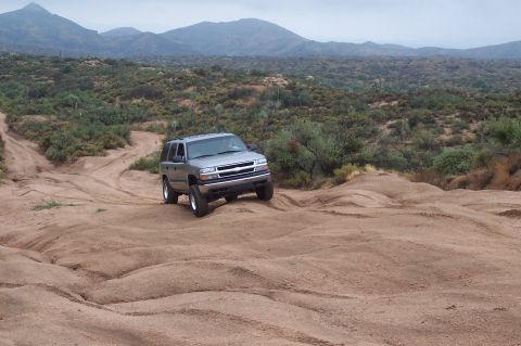1992 Ford Bronco - This silver SUV navigates a dusty, off-road trail with arid hills and mountains in the distance.