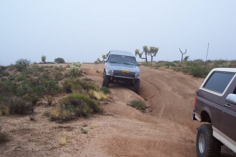 1992 Ford Bronco - Two SUVs navigate a dusty trail in a desert environment with sparse vegetation and a hazy sky.