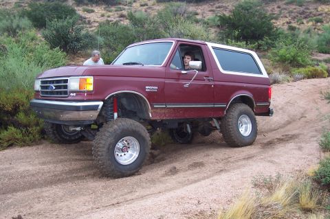 1992 Ford Bronco - A maroon 1992 Ford Bronco is driven off-road on a dirt path with vegetation in the background.