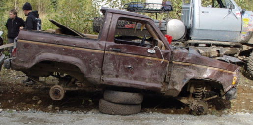 Stripped maroon pickup body sitting on bare wheels at a muddy trail with onlookers