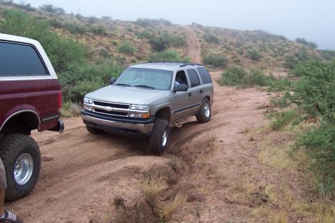 1992 Ford Bronco - Two SUVs navigate a dusty, off-road trail with sparse vegetation and hills in the background.