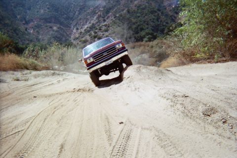 1990 Ford Bronco - dark red SUV crests a sandy berm head-on, front wheels lifting, on a rutted off-road trail in a brushy canyon.