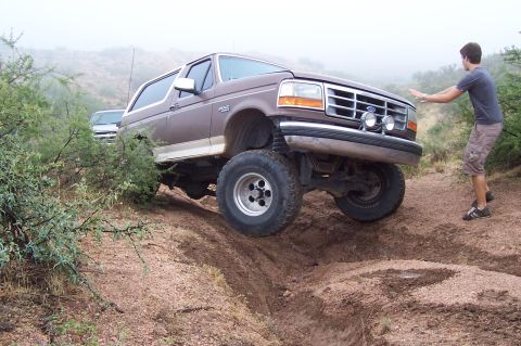 1992 Ford Bronco - A person pushes a lifted Ford Bronco as it navigates a steep dirt incline, with another vehicle visible in the background.