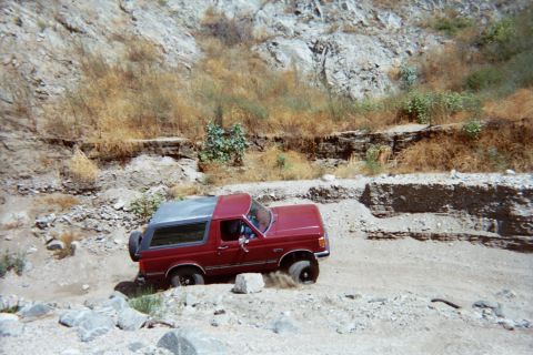 1990 Ford Bronco - red SUV with a black hardtop picks its way along a narrow dirt ledge beneath a steep rocky canyon wall.