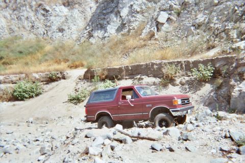 1990 Ford Bronco - maroon SUV sits among loose rocks at the foot of a rugged, sun-bleached canyon slope.