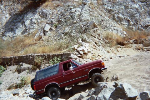 1990 Ford Bronco - maroon SUV with its window down eases down a steep, rocky off-road grade in a sandy canyon.