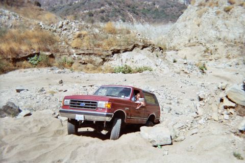 1990 Ford Bronco - maroon Ford SUV, its Ford grille badge visible, rests on rocky ground in a dry canyon with a person at the window.