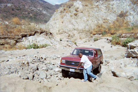 1990 Ford Bronco - man in a white shirt braces against the front of a red Ford SUV wedged on rocks in a dry, sandy canyon.