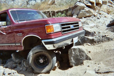 1990 Ford Bronco - Close-up of a maroon Ford SUV's chrome grille and Ford oval as its front tire climbs over a large rock.