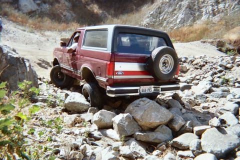 1990 Ford Bronco - rear of a red-and-white two-tone Ford SUV with a rear-mounted spare crawls over a boulder-strewn trail.