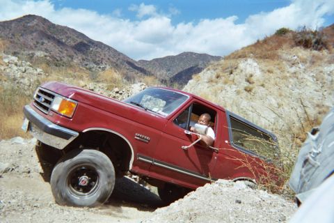 1990 Ford Bronco - maroon Ford SUV leans at an angle on a rocky off-road obstacle, driver visible, with dry mountains beyond.