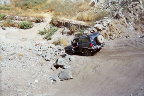 1990 Ford Bronco - dark gray SUV with a rear-mounted spare sits on a sandy off-road trail beside scattered boulders in a canyon.