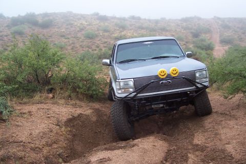 1992 Ford Bronco - This silver SUV navigates a challenging off-road path, equipped with a custom bumper and distinctive smiley face auxiliary lights.