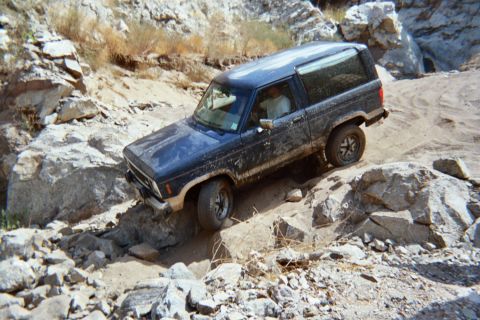 1990 Ford Bronco - mud-splattered dark blue compact SUV climbs over rocks on a steep off-road trail beneath a rocky slope.
