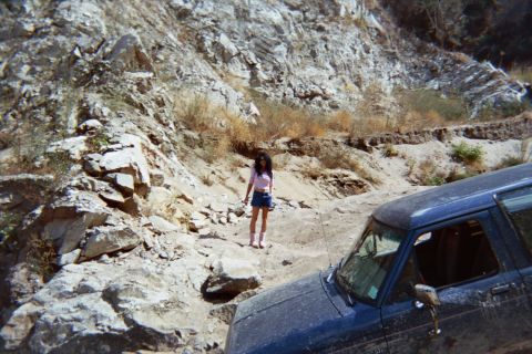 1990 Ford Bronco - young woman in shorts stands on rocky ground next to a dust-covered dark SUV in a rugged canyon.