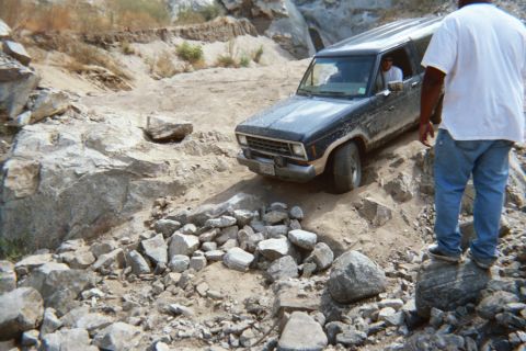 1990 Ford Bronco - mud-covered dark SUV crawls over loose rock as a man in a white shirt watches from the right in a dry canyon.