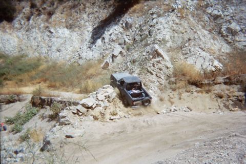 1990 Ford Bronco - dark open-top off-road vehicle powers up a steep sandy embankment below a rocky canyon wall.