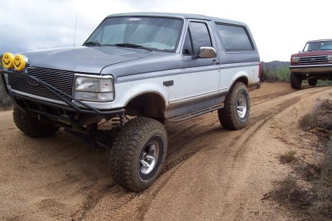 1992 Ford Bronco - Two Ford Broncos navigate a dusty off-road path, one equipped with auxiliary lights.