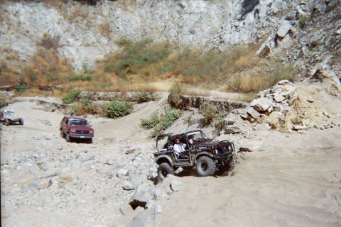 1990 Ford Bronco - black open-top off-roader crawls over rocks near a parked red SUV and other vehicles in a sandy quarry-like canyon.