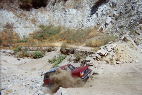 1990 Ford Bronco - maroon SUV throws up a cloud of dust while climbing a sandy off-road trail below a rocky canyon wall.