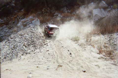 1990 Ford Bronco - red SUV powers up a rocky off-road slope, half-hidden in a billowing cloud of dust.