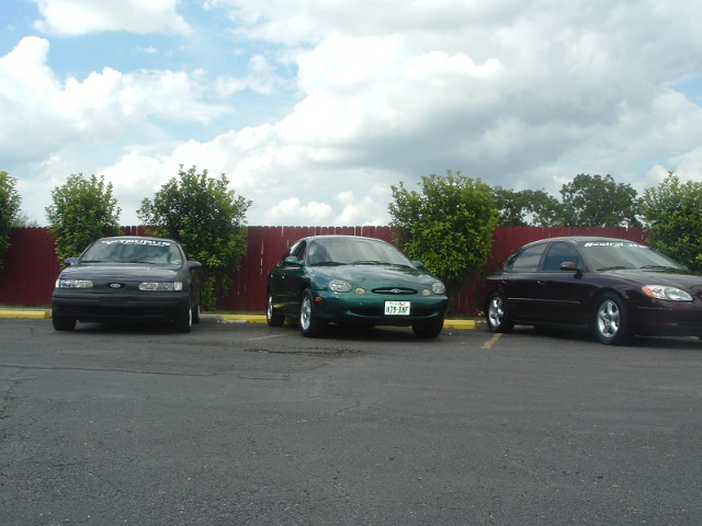 Three Ford Taurus sedans parked in a lot under a cloudy sky. The middle car is green, the left is dark, and the right is maroon