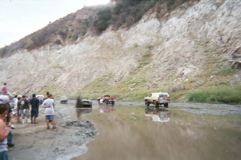 1990 Ford Bronco - crowd of spectators watches several off-road trucks and SUVs splash through a muddy water hole below a tall canyon wall.