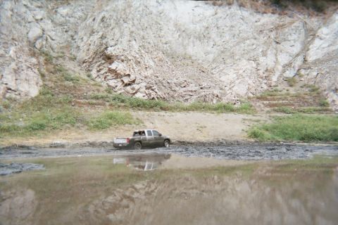 1990 Ford Bronco - pickup truck wades through a muddy water crossing, reflected in the water, at the base of a steep rocky canyon wall.