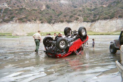 1990 Ford Bronco - red SUV lies upside down on its roof in a muddy pit while several people, including a uniformed officer, gather around.
