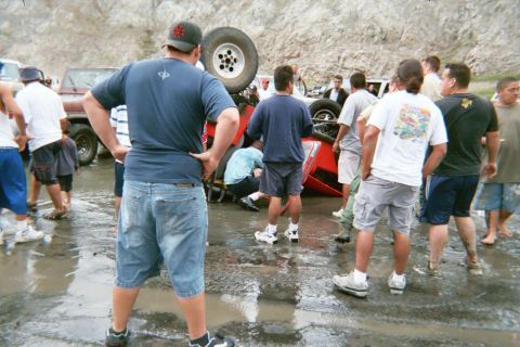 1990 Ford Bronco - crowd of onlookers stands around an overturned red vehicle sitting wheels-up in a muddy clearing.