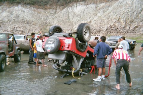 1990 Ford Bronco - red Jeep Wrangler rests overturned in a shallow muddy stream as a crowd of onlookers gathers near other off-road trucks.