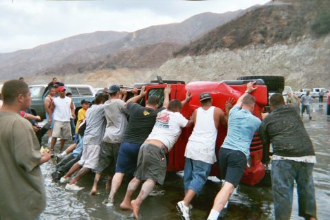 1990 Ford Bronco - line of people heave together to flip an overturned red Jeep back upright in a shallow stream below barren hills.