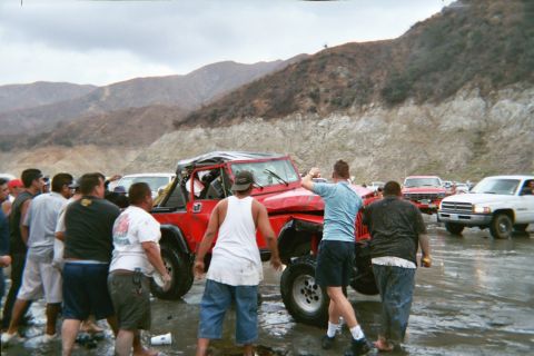 1990 Ford Bronco - Onlookers and trucks surround a battered red Jeep being maneuvered upright in a muddy off-road riverbed.