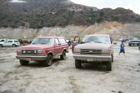 1990 Ford Bronco - red Ford Bronco and a red Ford pickup sit side by side on a dusty lot backed by rocky hillsides.