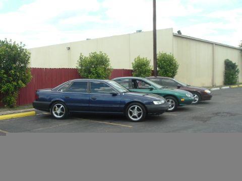 1993 Ford Taurus - Three sedans are parked in a line next to a building with a red fence in front.