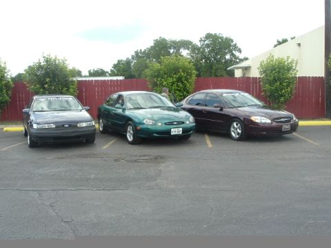 1993 Ford Taurus - Three Ford Taurus sedans are parked in a lot, with a red fence and trees in the background.