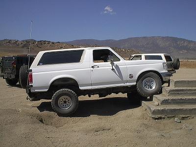 1990 Ford Bronco - lifted white Ford Bronco on oversized tires sits in profile on a dusty lot with mountains in the background.