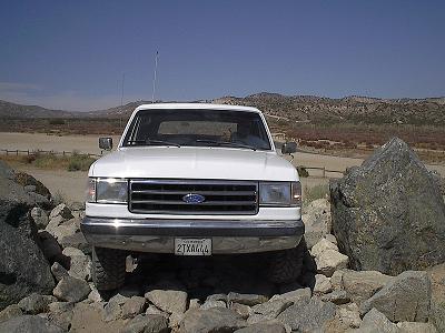 1990 Ford Bronco - white Ford truck with chrome grille is perched atop a rock pile on a desert flat under a clear blue sky.