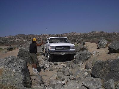 1990 Ford Bronco - man in a yellow cap spots a white Ford truck as it negotiates a rocky section of a desert trail.