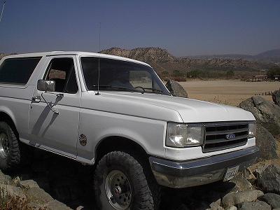 1990 Ford Bronco - white Ford Bronco with hardtop and chrome grille sits on rocks in an open desert field near a fence and trees.