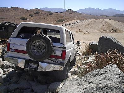 1990 Ford Bronco - rear of a white Ford Bronco with red stripe and spare tire climbs over boulders at a busy off-road riding area.