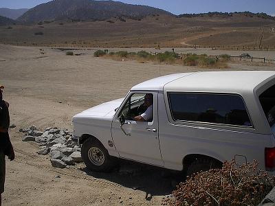 1990 Ford Bronco - white Ford Bronco with a person at the wheel sits beside a small rock pile on a wide desert flat with distant hills.