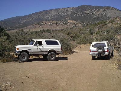 1990 Ford Bronco - white Ford Bronco and a second light-colored SUV sit on a dirt trail framed by brush and a forested mountain.