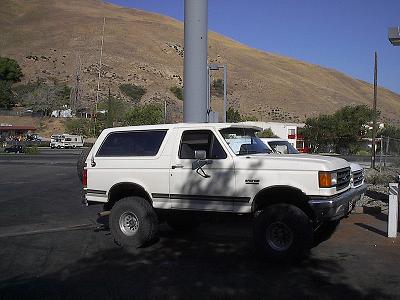 1990 Ford Bronco - lifted white Ford Bronco on large tires is parked at a gas station with bare brown hills rising in the background.