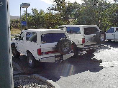 1990 Ford Bronco - Two white Ford Bronco SUVs with rear-mounted spares are parked one behind the other in a paved lot near shrubs.