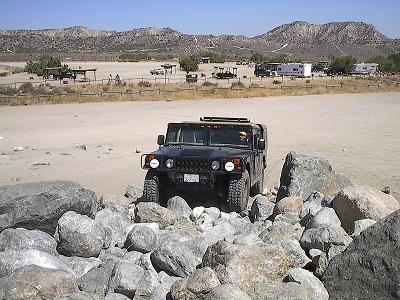 1990 Ford Bronco - black Hummer H1 climbs over a pile of large gray boulders at a desert off-road riding area with camps in the distance.