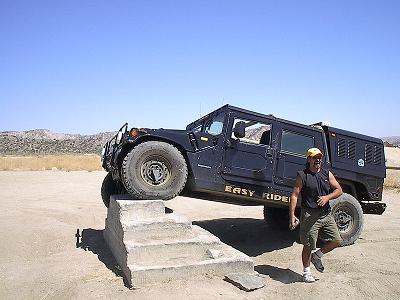 1990 Ford Bronco - man poses beside a black 'EASY RIDER' Hummer H1 that is perched on a concrete step ramp in a dirt clearing.