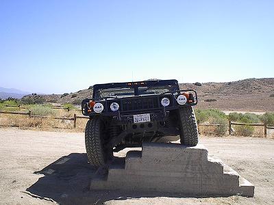 1990 Ford Bronco - black Hummer H1 articulates over a concrete step obstacle, one front wheel lifted, in a desert pullout.